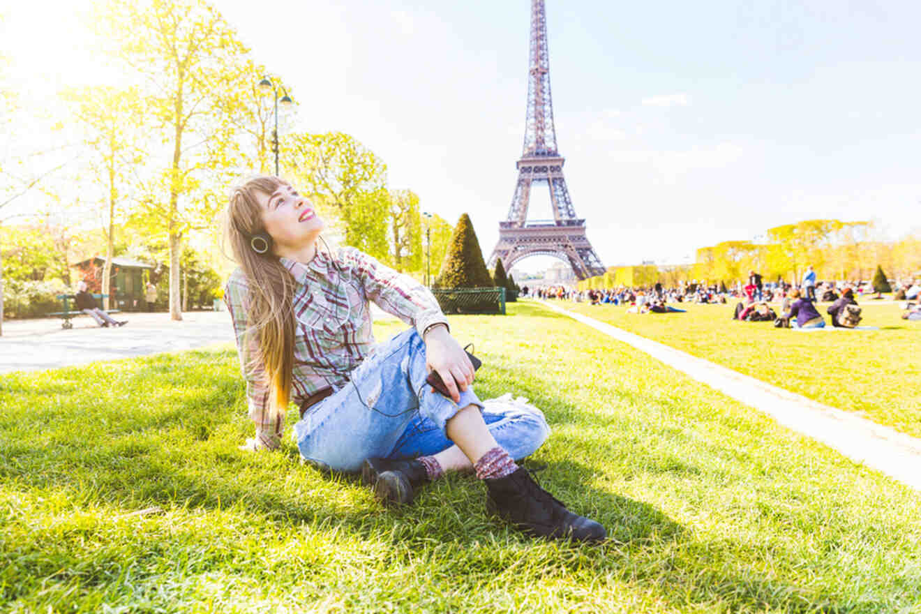 A person sitting on the grass in front of the Eiffel Tower on a sunny day, looking up and smiling, with several other people scattered around enjoying the open space.