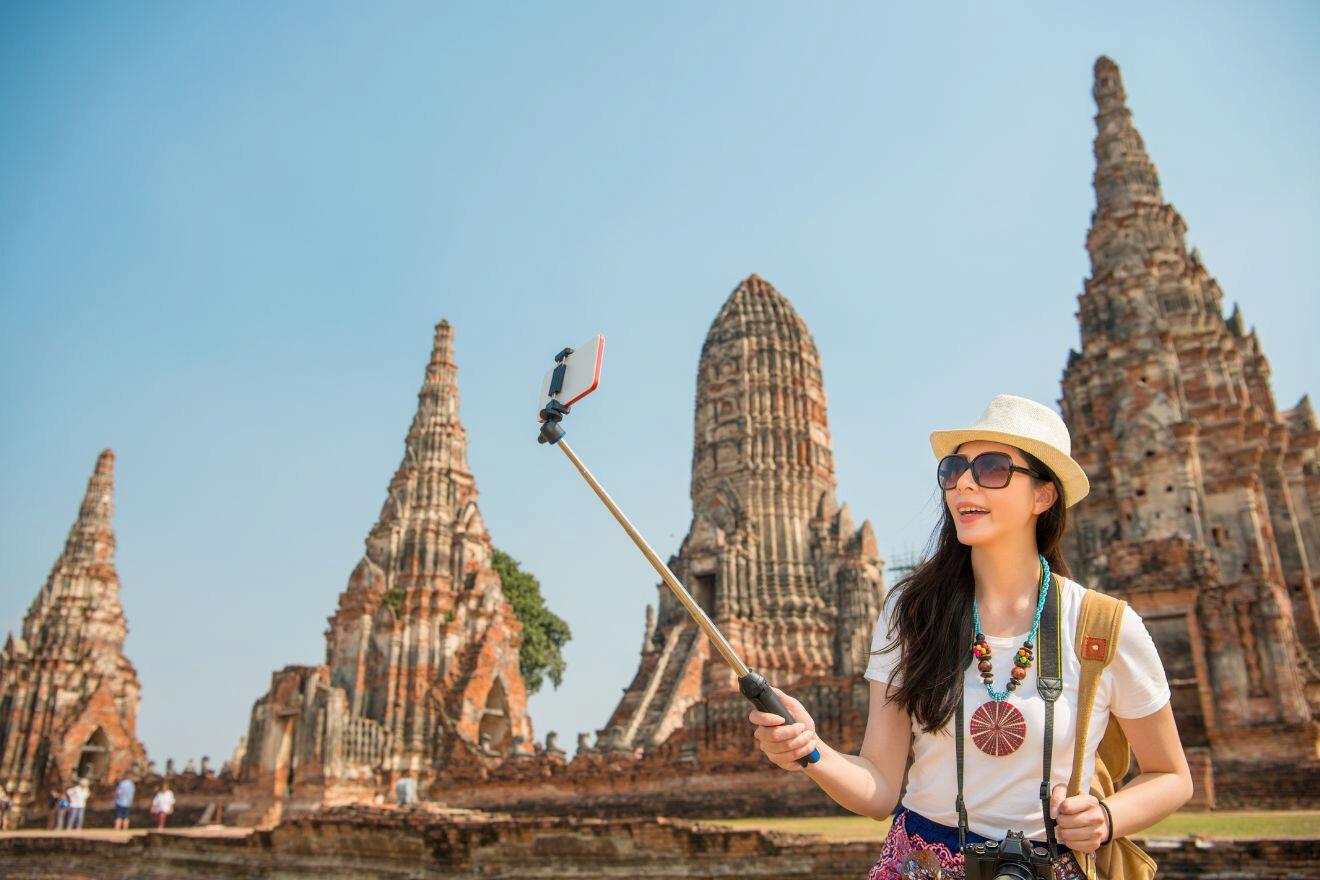 A woman in a hat and sunglasses takes a selfie with a selfie stick in front of ancient temple ruins under a clear blue sky.