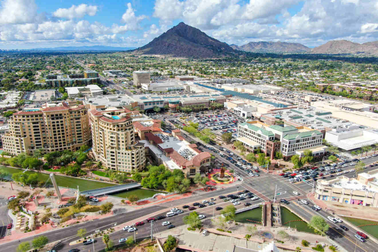 Aerial view of an urban area with a busy intersection, several mid-rise buildings, parking lots, and a mountainous backdrop under a partly cloudy sky.