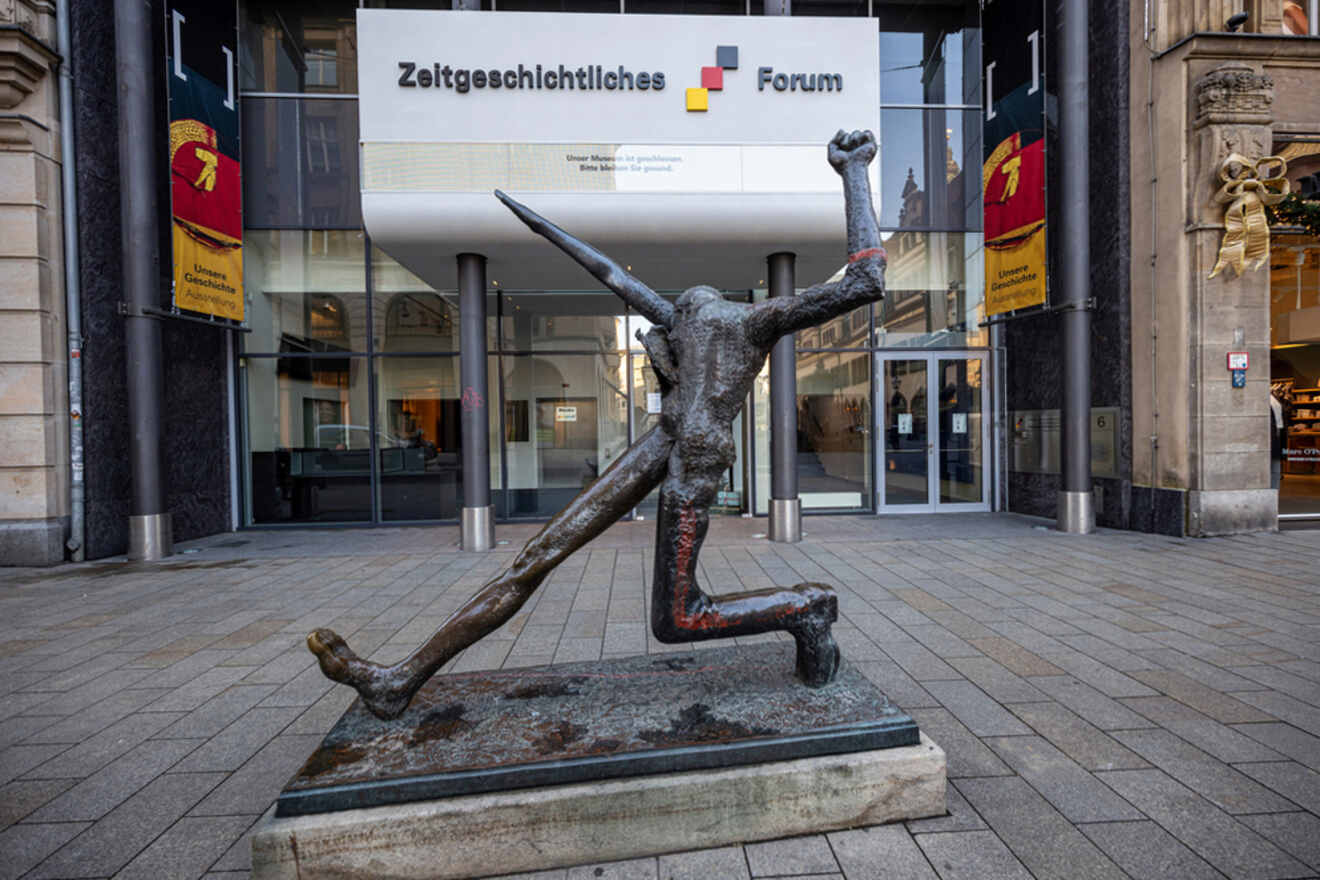 A bronze statue of a figure in a dynamic pose with one arm raised and the other holding a spear, located in front of the Zeitgeschichtliches Forum building.