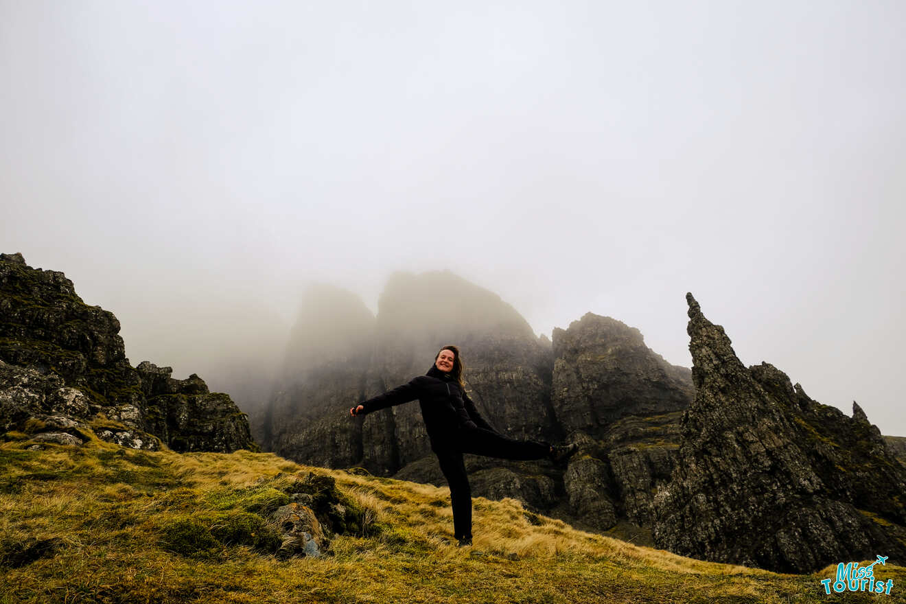 Author of the post standing on a grassy slope, striking a playful pose with one leg lifted, surrounded by rocky formations and misty, cloud-covered peaks.