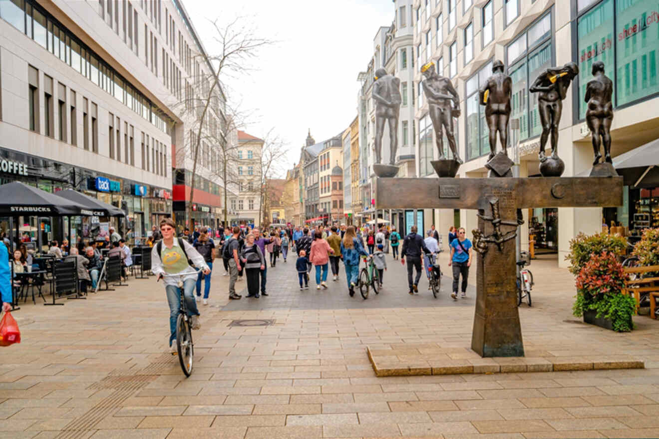 A busy pedestrian street featuring people walking and biking, lined with shops and cafes on both sides, with a modern sculpture in the foreground.