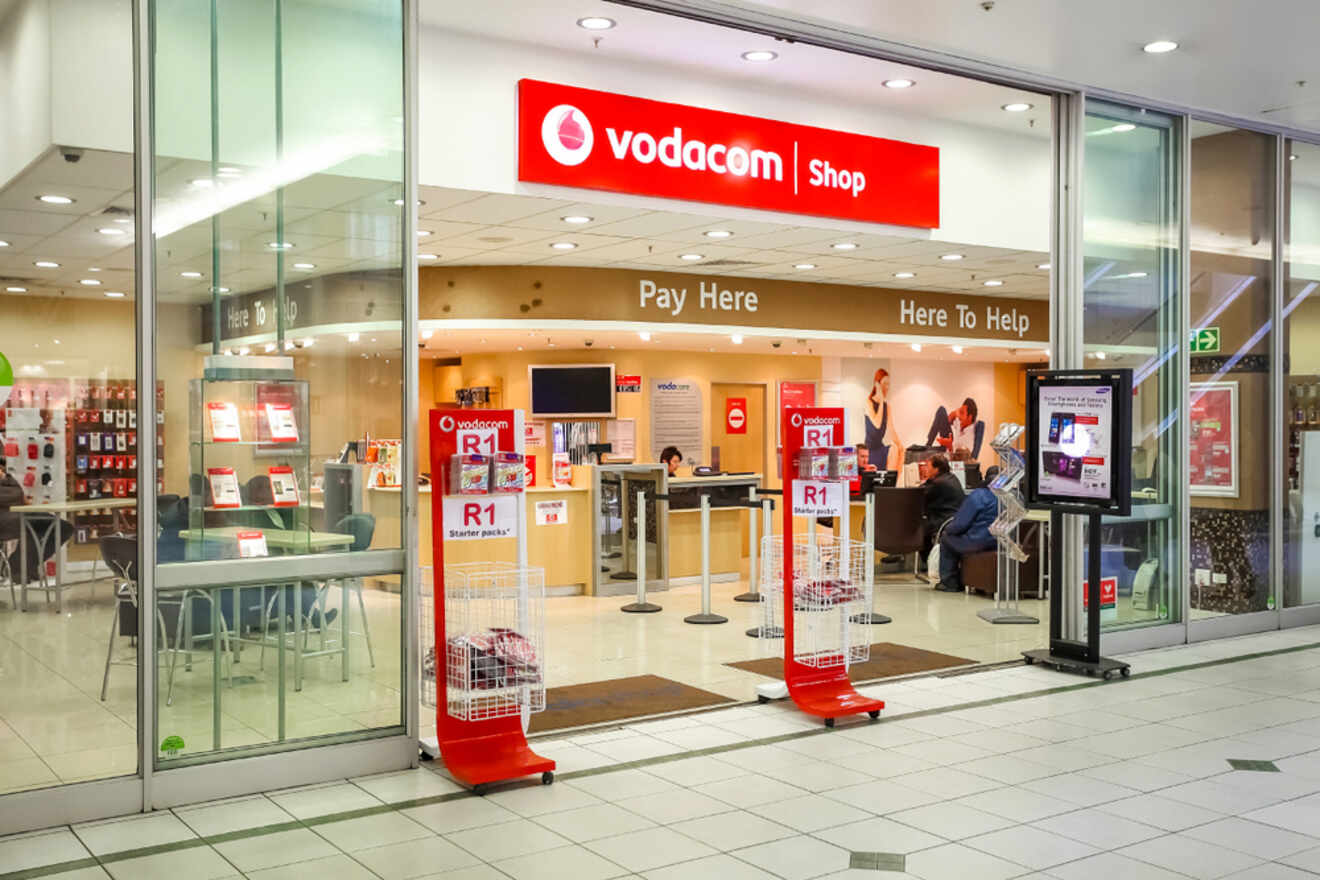 A Vodacom store in a shopping mall with a glass entrance, promotional displays, and signs stating "Pay Here" and "Here to Help." Customers are seen interacting with staff inside.