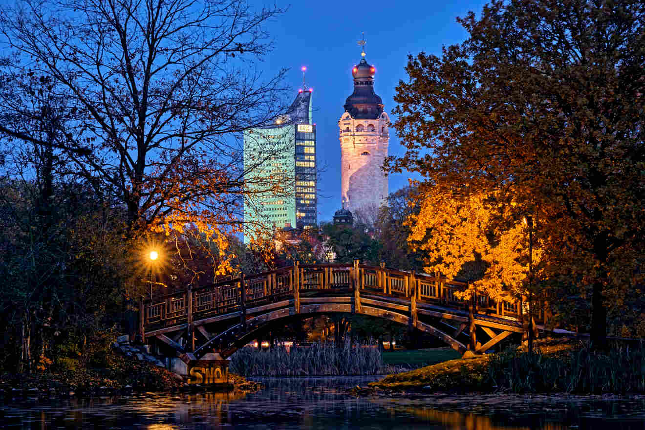 A wooden bridge over a pond is framed by trees with two tall buildings illuminated in the background, one modern and one historic, during twilight.