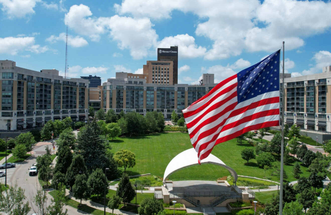 A large American flag flying in front of an urban park with a performance pavilion and surrounding buildings.