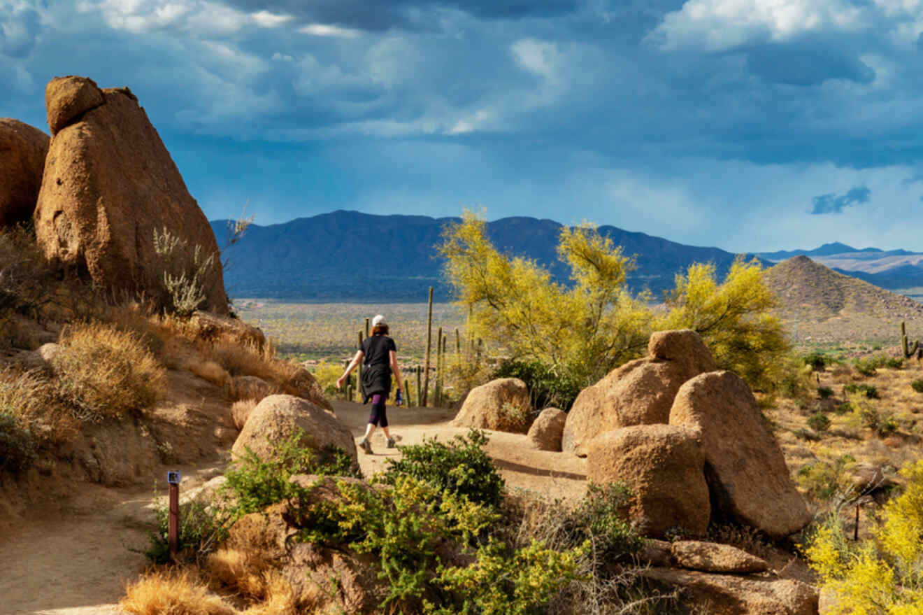A hiker walking along a desert trail surrounded by large boulders and cacti, with mountains in the background