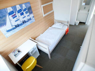 A small, modern bedroom with a single bed, a desk with a yellow chair, a wall-mounted sailing poster, and a white sink area in the corner.