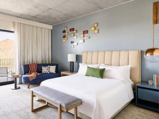 A modern bedroom with a beige headboard, white bedding, a blue sofa, and colorful wall art.