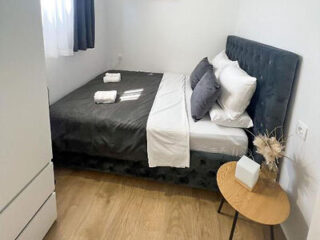 Uma cama impecavelmente arrumada, com roupa de cama escura, lençóis brancos e duas toalhas dobradas, está situada em um pequeno quarto com piso de madeira, uma mesa de cabeceira e uma janela com cortinas translúcidas e escuras.