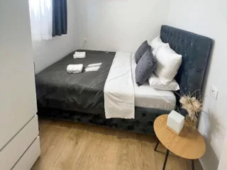 A neatly made bed with dark bedding, white sheets, and two folded towels sits in a small room with wooden flooring, a side table, and a window with sheer and dark curtains.