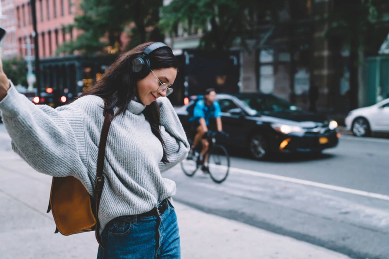 A person wearing headphones and a gray sweater walks down a city street with cars and a cyclist in the background. They have a brown shoulder bag and appear to be enjoying music or a phone call.