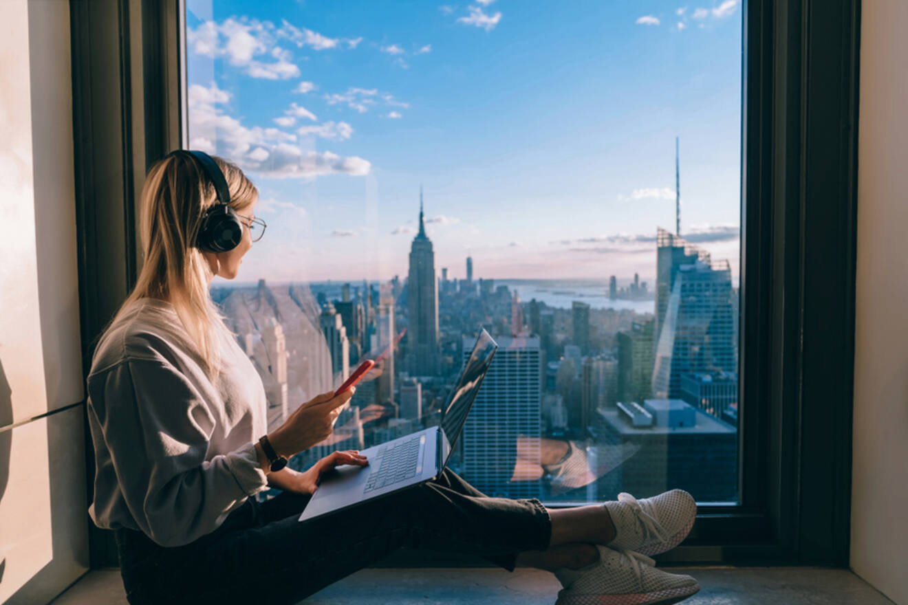 A person wearing headphones sits by a large window with a laptop and pen, overlooking a cityscape with tall buildings during the day.