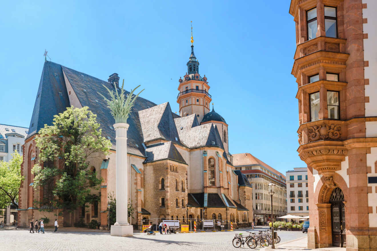 A historic church with a tall, ornate column stands in a sunny square. Nearby buildings and a few bicycles are visible.