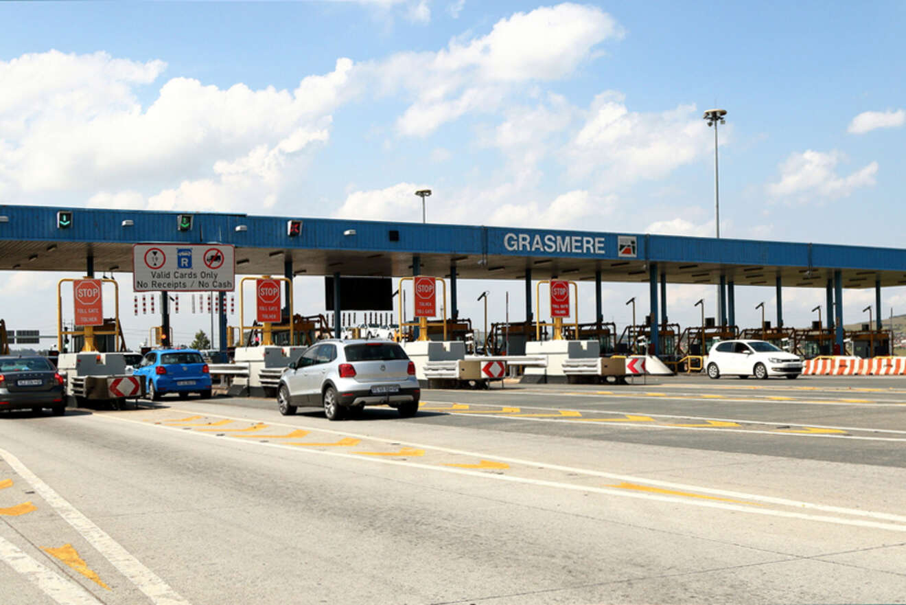 Cars passing through a toll plaza under a blue sky with white clouds. The sign above the plaza reads "GRASMERE" and has various smaller signs with instructions for drivers.