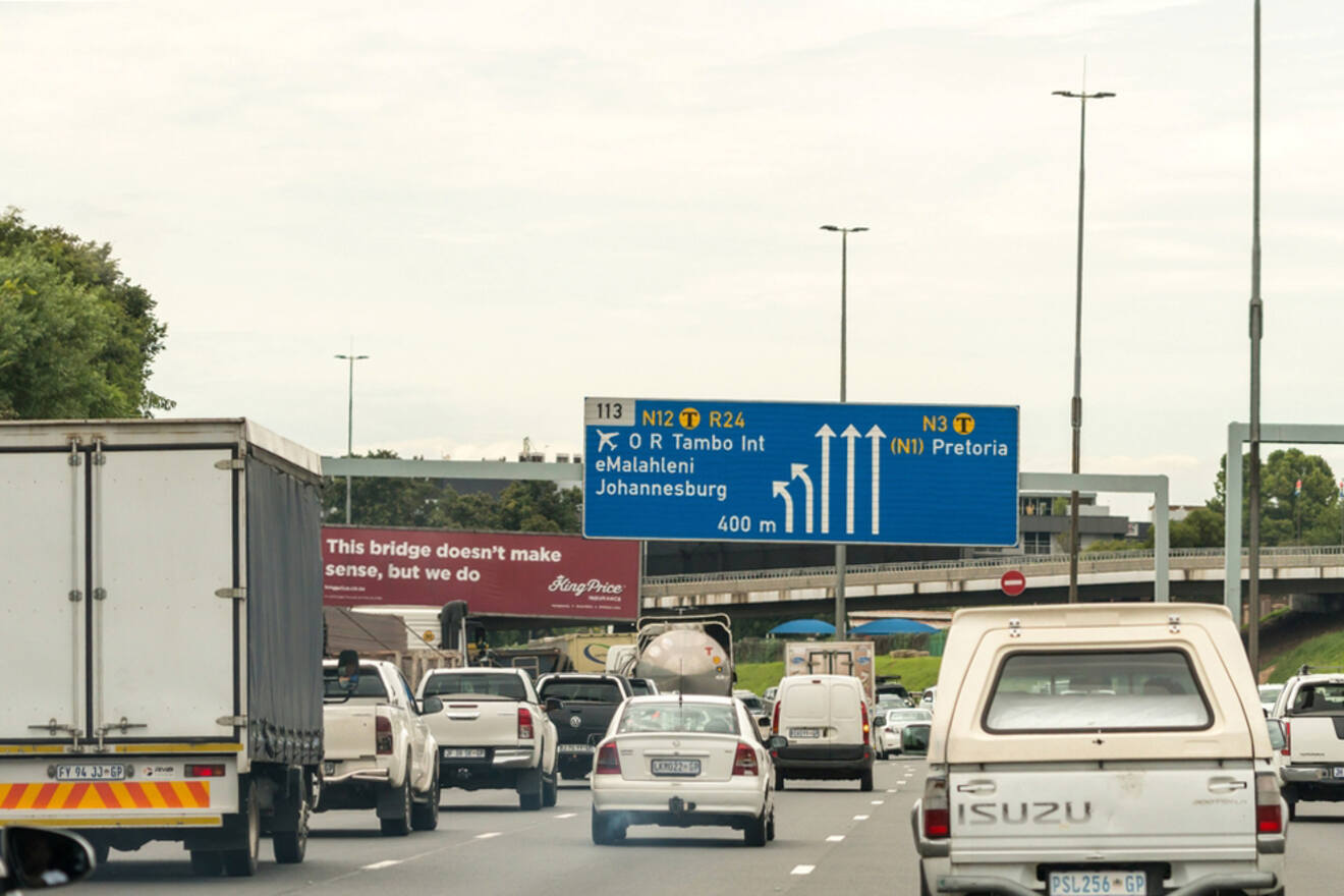Cars driving on a highway towards Johannesburg and Pretoria, with a road sign indicating directions and a billboard reading "This bridge doesn’t make sense, but we do" visible on the left.