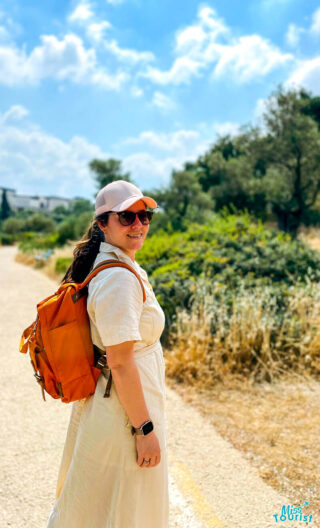 Author of the post in a white dress and orange backpack smiles while walking on a sunny path, wearing a pink cap and sunglasses with greenery in the background.