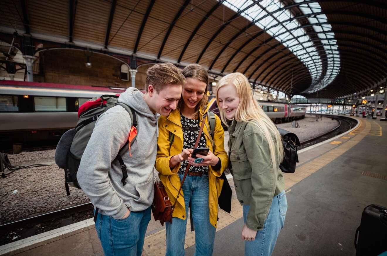 Three people stand on a train platform under a curved roof, looking at a smartphone together and smiling.