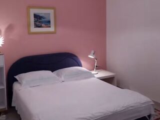 Uma cama impecavelmente arrumada com lençóis brancos, dois travesseiros, uma cabeceira azul, uma mesa de cabeceira com um abajur e um quadro emoldurado em uma parede rosa.