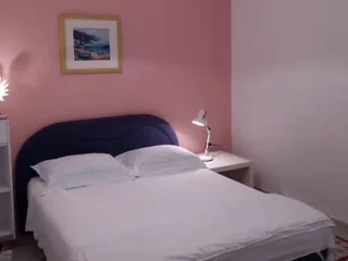 A neatly made bed with white linens, two pillows, a blue headboard, a bedside table with a lamp, and a framed picture on a pink wall.