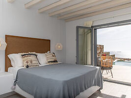 A bedroom with a large bed covered in grey bedding, leading to a balcony with a view of the sea.