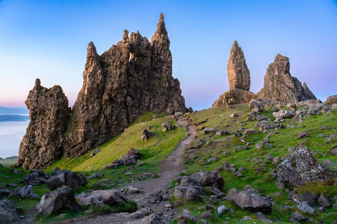 A rocky landscape with tall spires and a dirt path winding through, set against a clear blue sky during early morning or late evening.