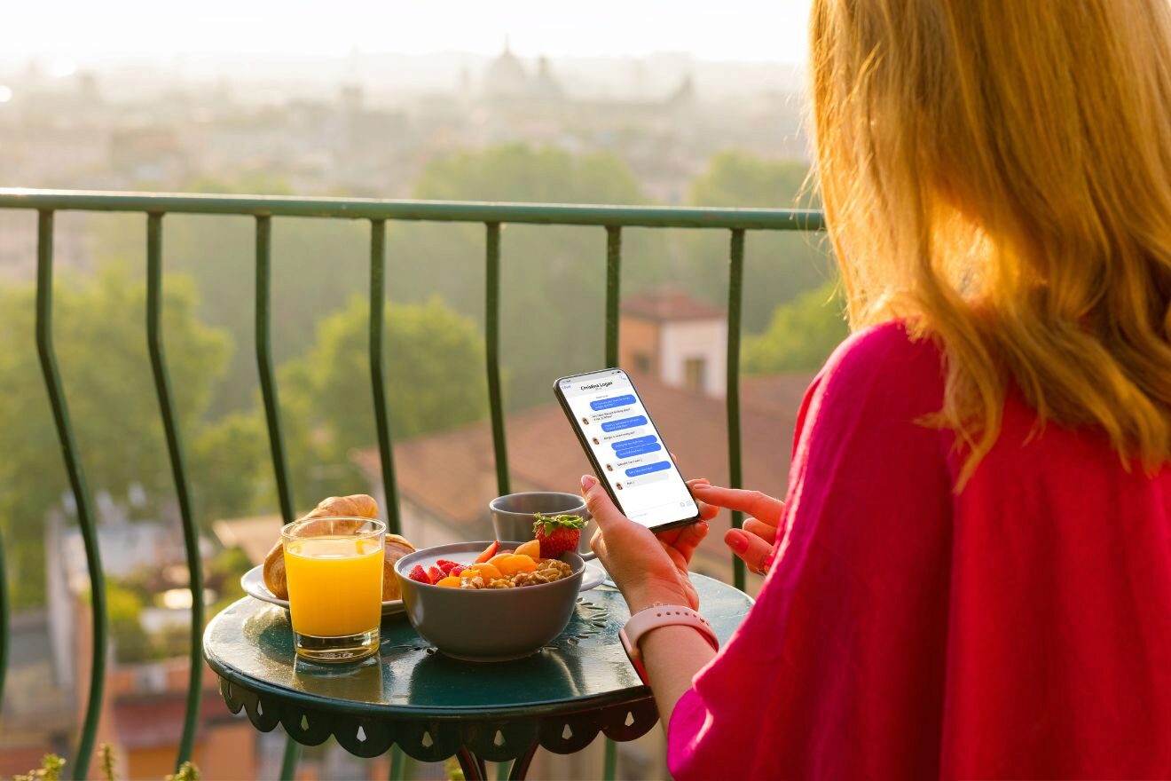 A person in a pink shirt is checking messages on a smartphone while sitting at a balcony table with breakfast items, including a glass of orange juice and a bowl of fruit, overlooking a cityscape.