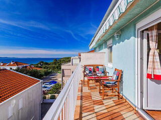 A balcony with wooden chairs and colorful cushions overlooks a coastal town and the ocean under a clear blue sky.