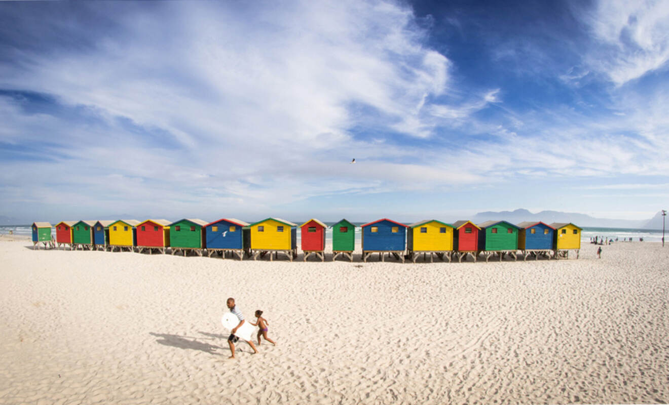 A row of colorful beach huts on a sandy beach under a blue sky. Two people walk across the sand, one carrying a surfboard.