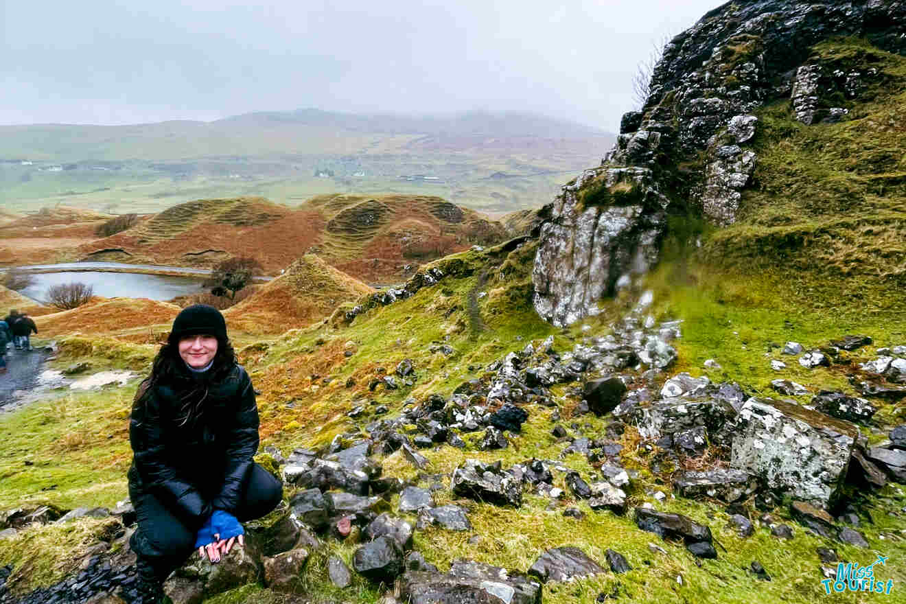 Author of the post in a black jacket and hat kneels on a rocky path amidst a green, hilly landscape on a cloudy day.