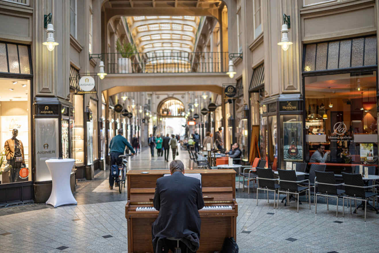 A person playing an upright piano in a shopping arcade with passersby and shops in the background. The arcade has a glass ceiling, creating a bright atmosphere.