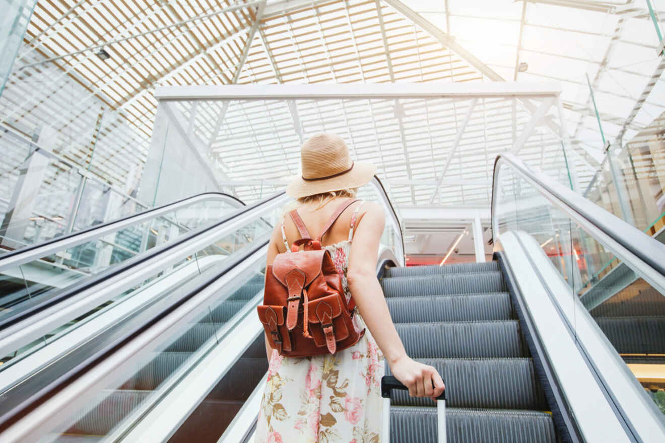 A person in a hat and floral dress rides an escalator in a modern, glass-ceilinged building, carrying a suitcase and wearing a brown backpack.