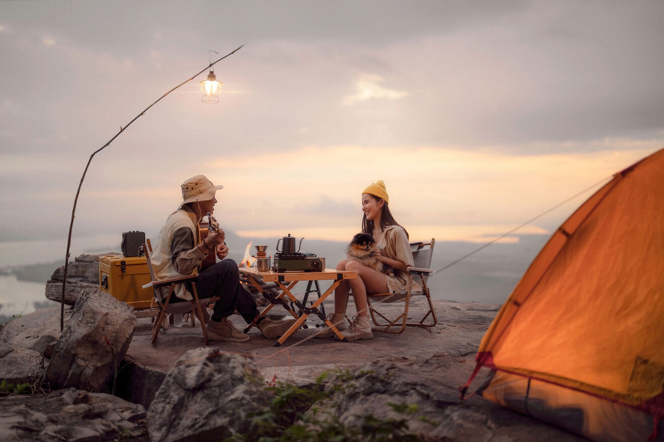 A man and woman sit by a camping setup on a cliffside at sunset, playing music and holding a dog. A tent is pitched nearby and a lantern hangs overhead.