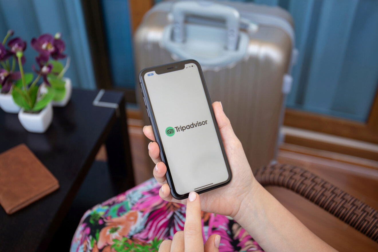 A person holding a smartphone displaying the Tripadvisor app in front of a silver suitcase and small plants.