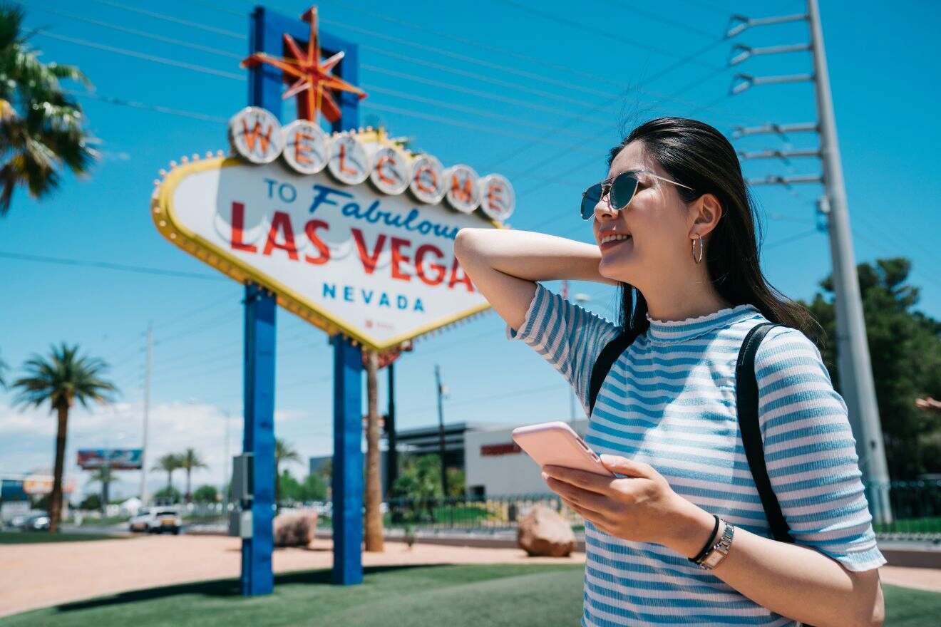 A person stands smiling in front of the "Welcome to Fabulous Las Vegas, Nevada" sign, holding a smartphone and wearing sunglasses and a striped shirt.