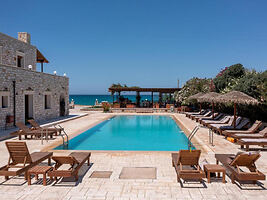 A spacious pool area with sun loungers, surrounded by a stone building and greenery, with the sea in the background.