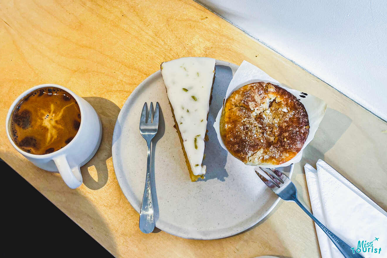 A cup of coffee sits next to a plate with a slice of cheesecake and a muffin on a wooden table. Two forks and napkins are placed beside the plate.