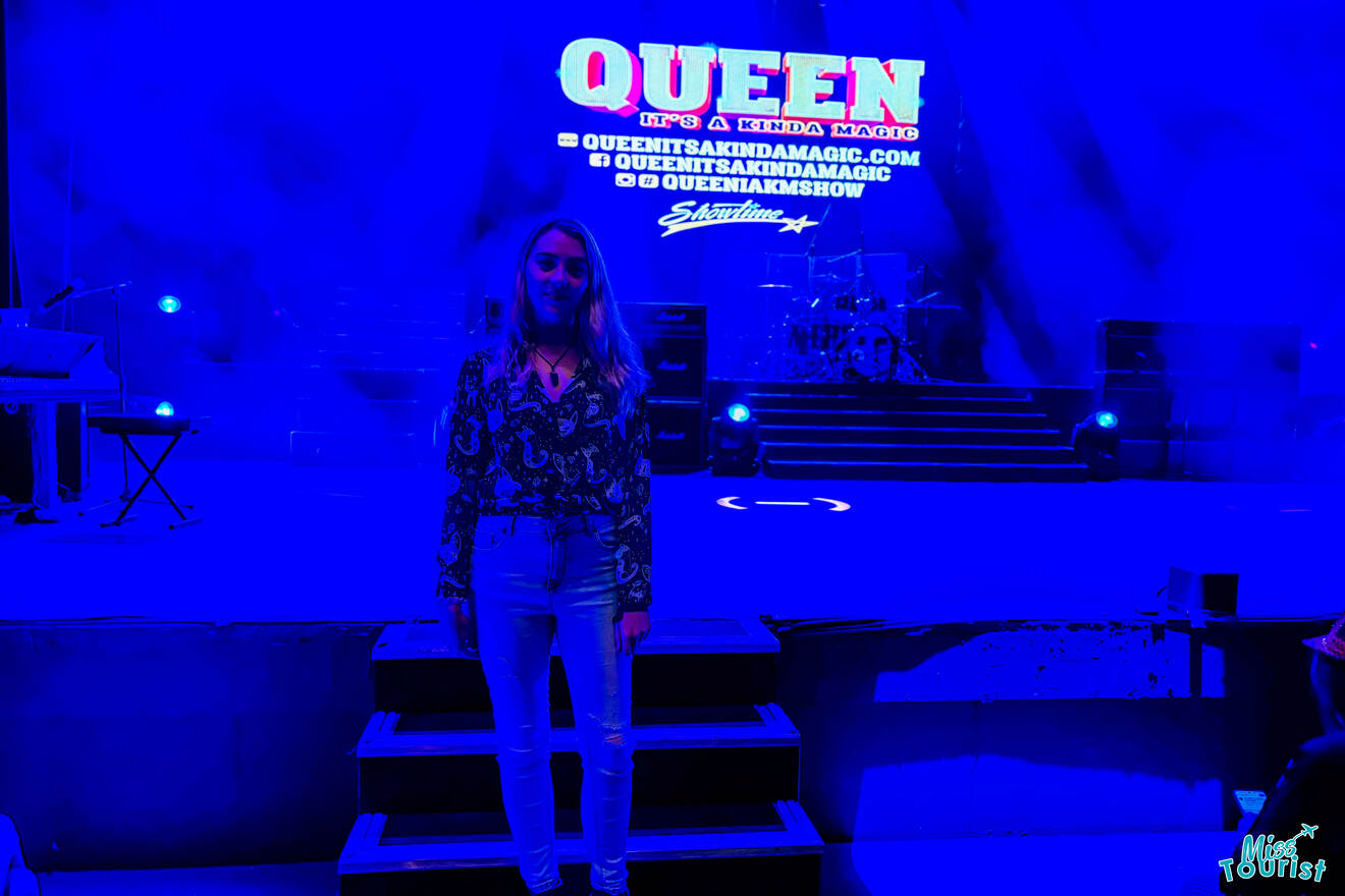 author of the post stands on a stage with a "Queen: It's a Kinda Magic" sign in the background. The stage is lit with blue lighting.