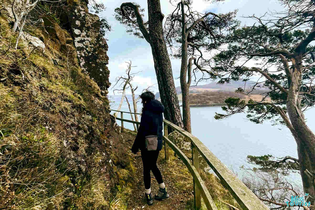 Author of the post wearing a jacket and backpack walks along a narrow, fenced path on a cliffside overlooking a body of water and trees.