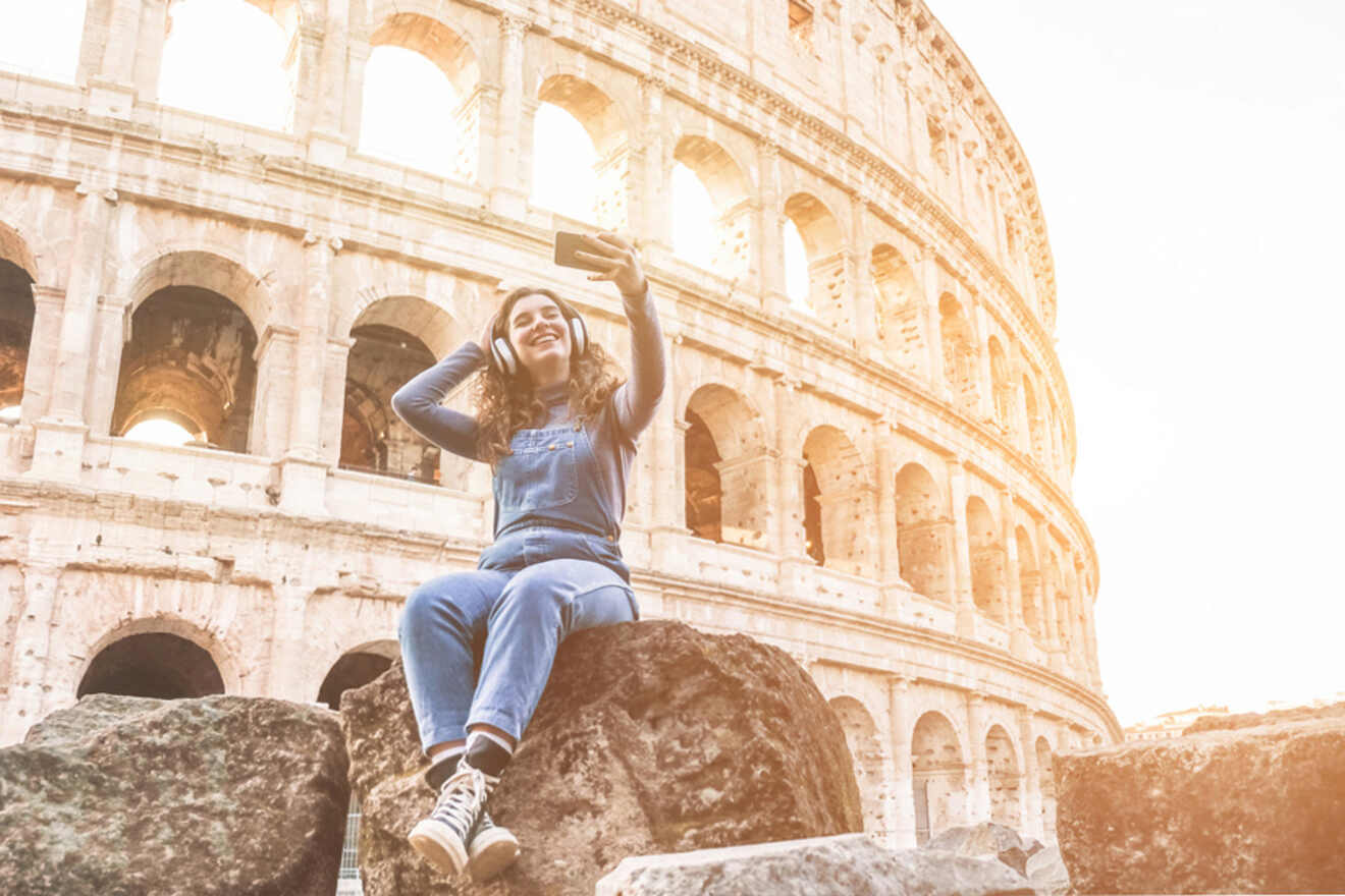 A person in a blue outfit is sitting on a rock, taking a selfie in front of the Colosseum.
