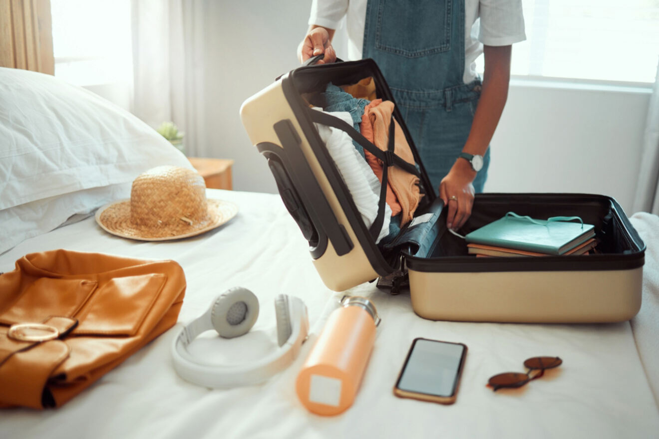 Person packing a suitcase on a bed, with various items laid out including a hat, clothes, headphones, a water bottle, a smartphone, and sunglasses.