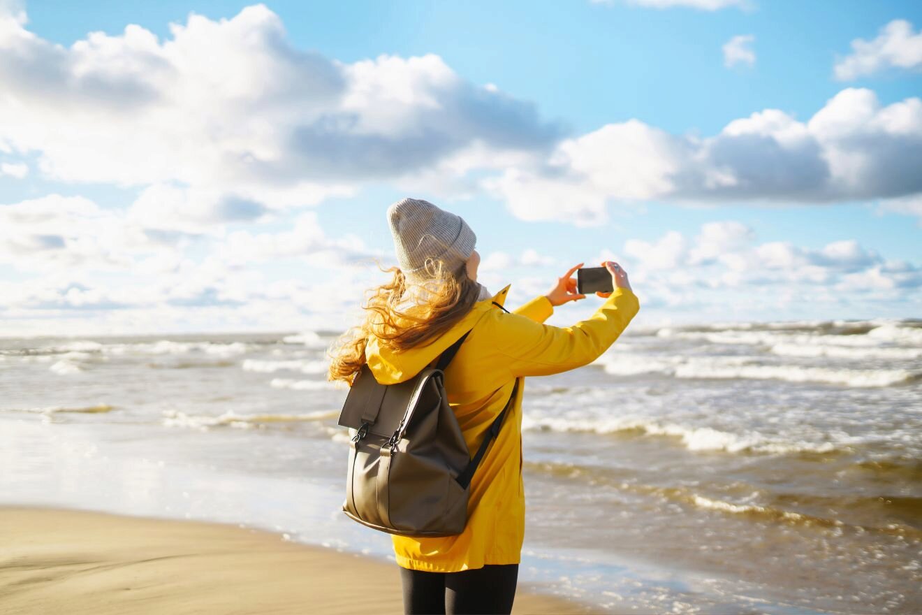 A person in a yellow jacket and gray beanie takes a photo with a smartphone on a beach with waves and a cloudy sky in the background.