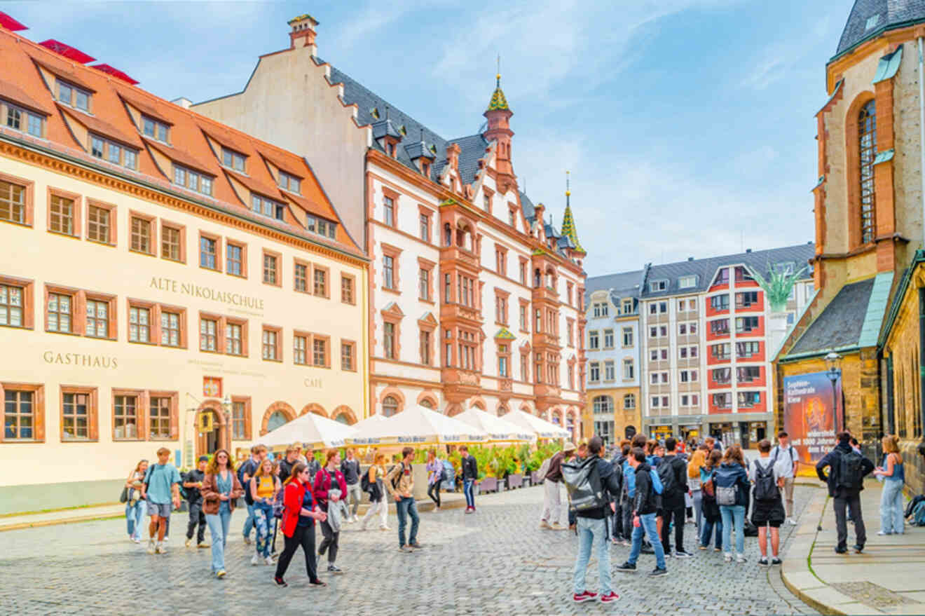 Crowd of people gather in a cobblestone plaza surrounded by historic European buildings with colorful facades and outdoor seating areas.