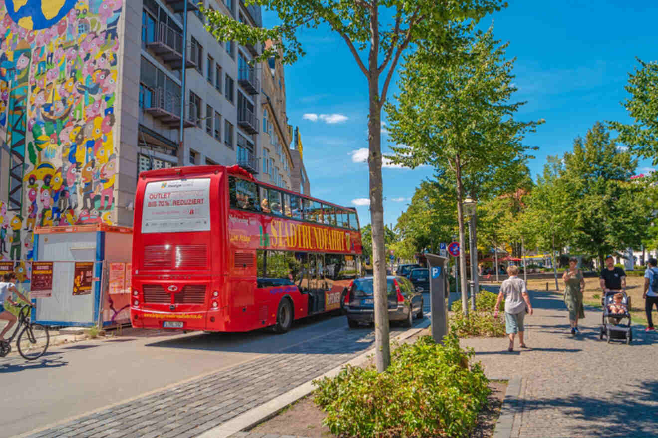 A red double-decker bus drives down a street lined with trees and colorful buildings while people walk and bike nearby.