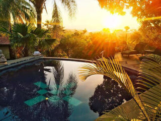 Sunset view over a serene swimming pool surrounded by tropical plants and trees, with lounge chairs on a wooden deck nearby.