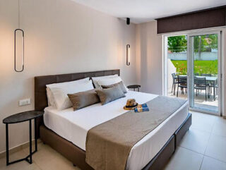 A modern bedroom with a large bed, neutral tones, and a patio door leading to an outdoor seating area.