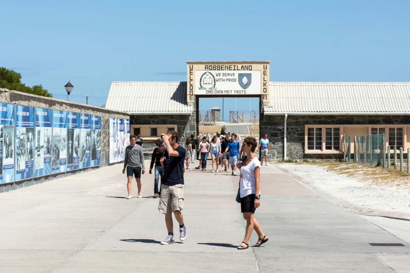 Visitors walking on a paved pathway towards the entrance of Robben Island, with informational displays on the left and a welcome sign overhead.