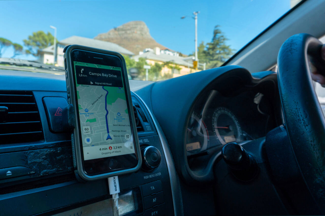 A smartphone with a navigation app is mounted on a car's dashboard, displaying directions to Camps Bay Drive. The driver's hand is on the steering wheel. Houses and a mountain are visible outside.