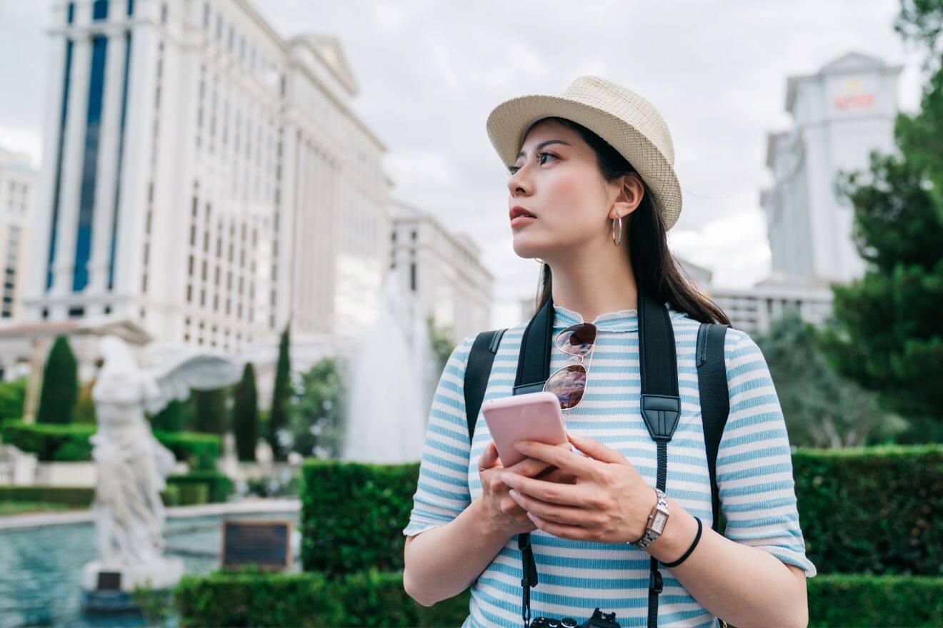 A woman in a hat and striped shirt holds a smartphone and looks around while standing outside near a fountain and large buildings.