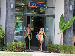 Two people, carrying surfboards, are exiting a building labeled "OCEANO." They are dressed in swimwear, and the entrance is surrounded by plants.