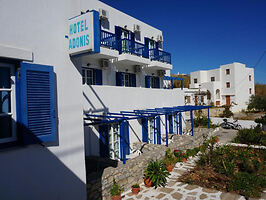 The exterior of Hotel Adonis featuring white walls, blue shutters, and balconies with blue railings, set against a bright blue sky.